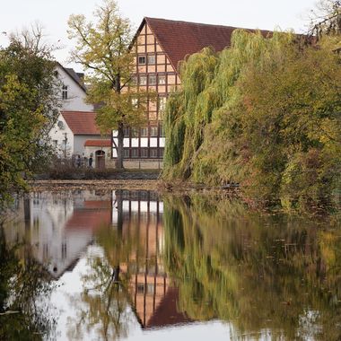 Ein Fachwerkhaus spiegelt sich in einem See, drumherum herbstliche Bäume.