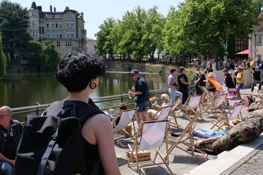 Menschen liegen auf Sonnenstühlen vor einem See in der Stadt, grüne Bäume im Hintergrund. Pride-Farben sind zu sehen auf kleinen Fähnchen.
