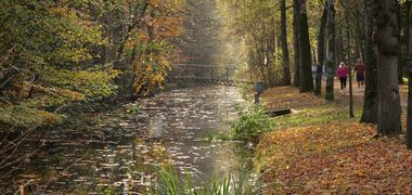Menschen laufen entlang eines Kanals umgeben von herbstlich eingefärbten Bäumen, im Hintergrund eine Brücke.