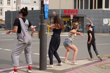 Vier Frauen tanzen an einer Straßenkreuzung vor der Ampel.