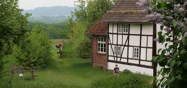 Neben einem Fachwerkhaus schweift der Blick auf grüne Koppeln und Hügel, Wanderer sitzen auf einer Bank und rasten.
