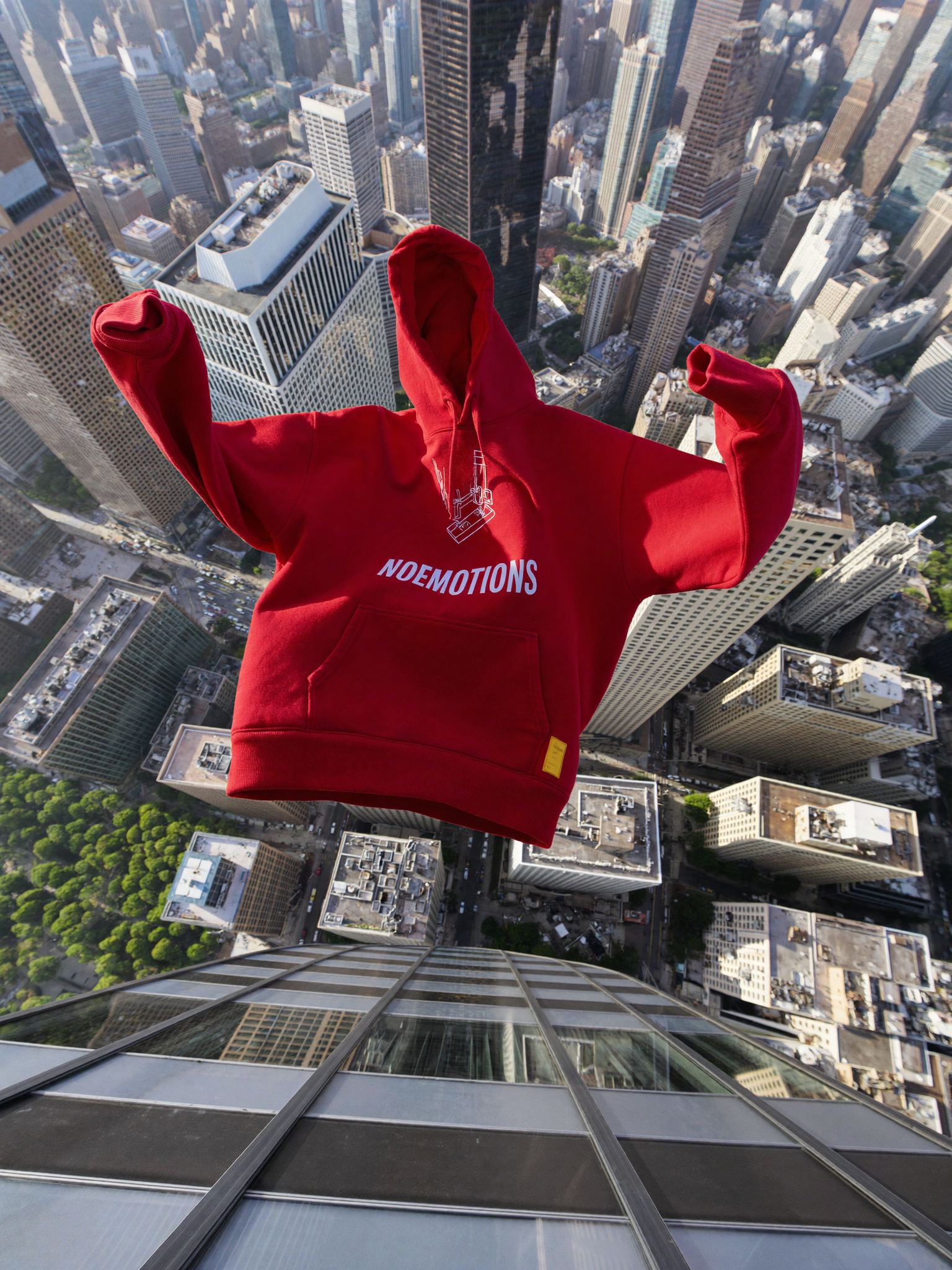 Red NOEMOTIONS hoodie flying through air above NYC skyscrapers, extreme aerial top-down view, sleeves spread wide like wings, sunlit city below, rooftops and streets in deep perspective, cinematic dramatic falling motion shot