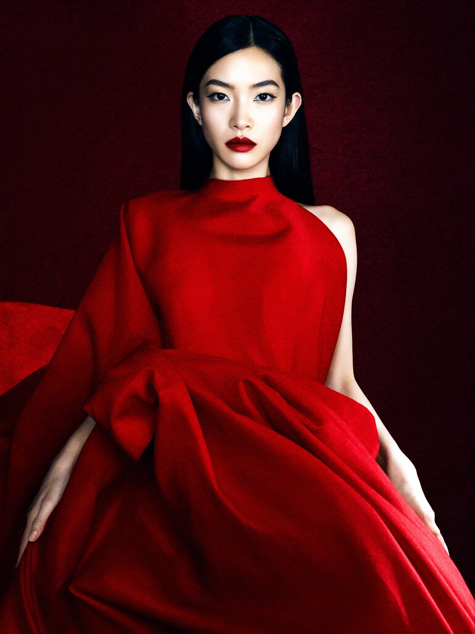 A jet-black-haired East Asian woman in a sculptural scarlet halter gown sits against a deep crimson backdrop, bold red lip, editorial fashion cover.