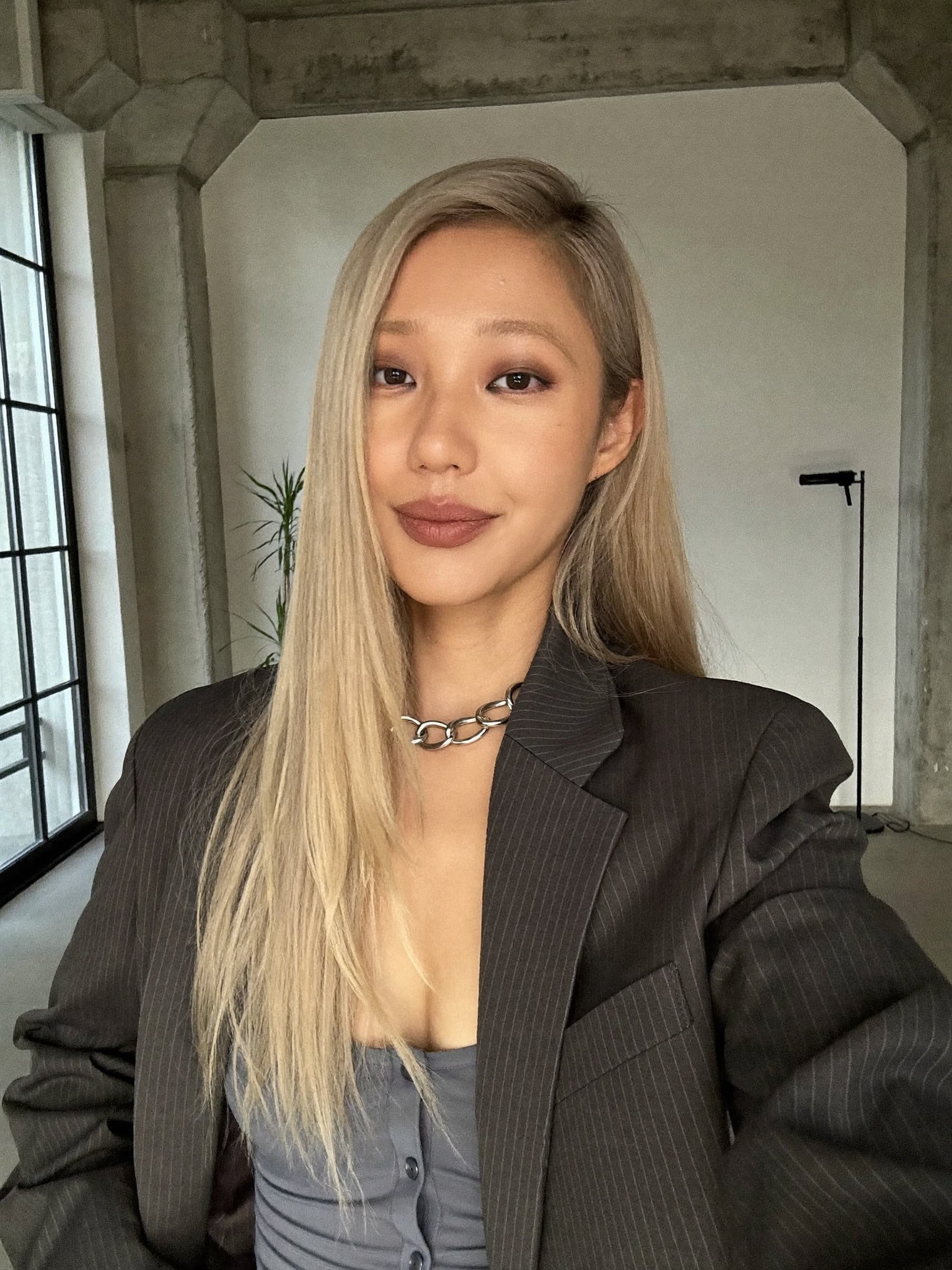 A platinum-blonde woman in an oversized pinstripe blazer and silver chain choker takes a soft-lit selfie in a concrete loft by a tall window.