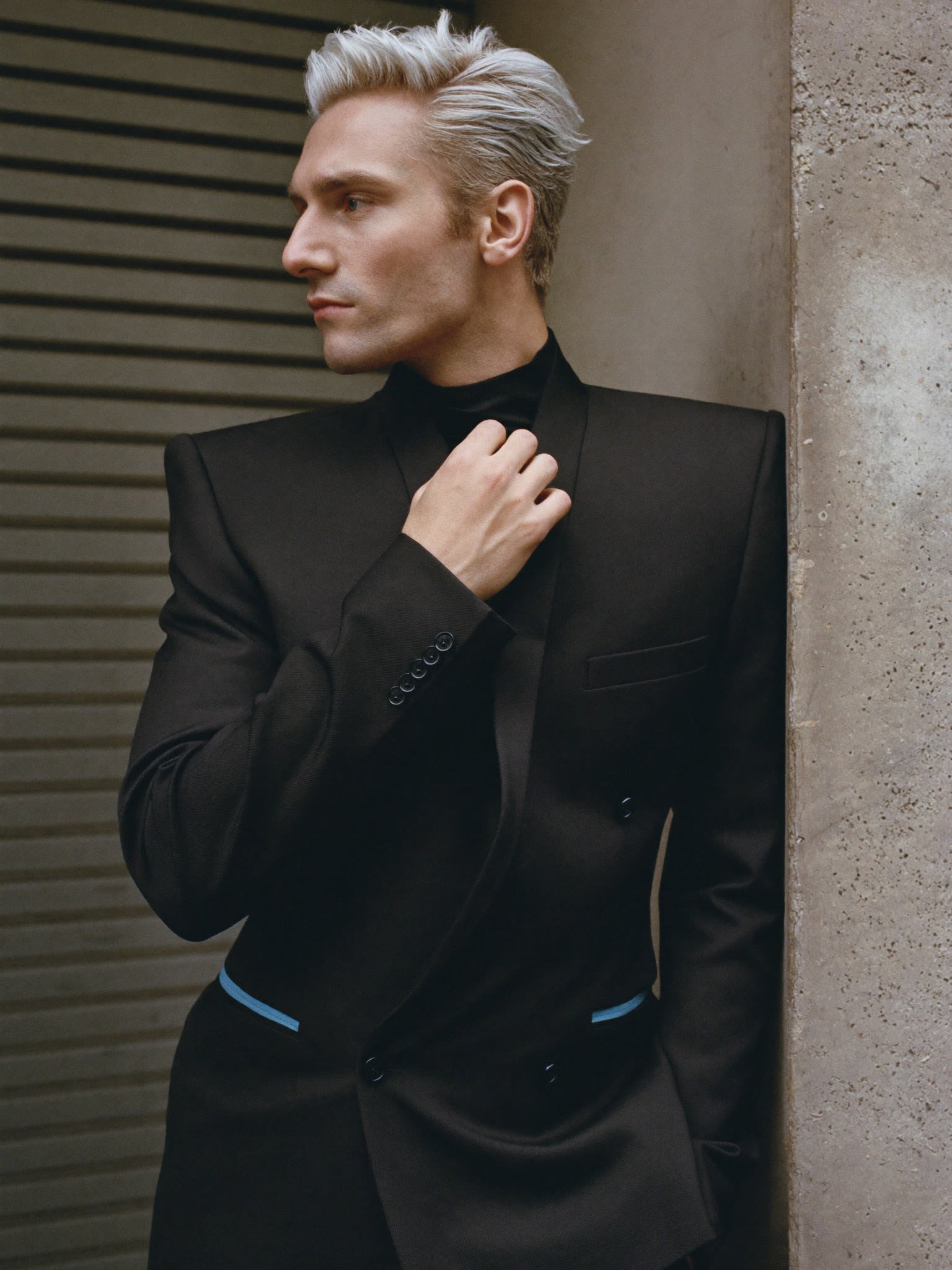 A platinum-haired clean-shaven man in a sharp black double-breasted blazer and turtleneck leans against a concrete pillar, glancing off-camera in profile.