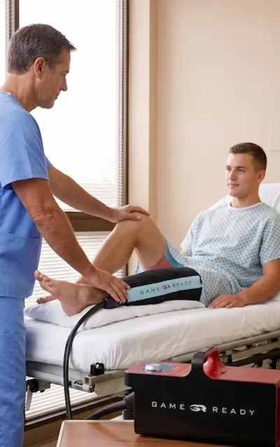 A young man is laying on an hospital bed with his leg wrapped in a Game Ready sleeve. A doctor is standing next to him.