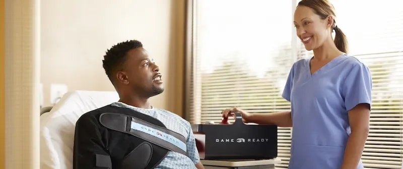 A patient in a hospital bed wears a shoulder wrap while a nurse in blue scrubs stands nearby with a Game Ready unit, smiling.