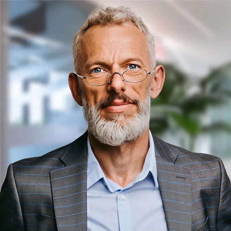 Board member in a gray blazer and light blue shirt with a soft office backdrop and a composed, friendly expression