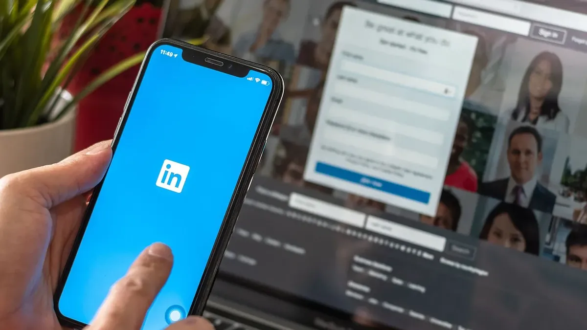Hand holding an iPhone with the LinkedIn app on the screen in front of a laptop displaying LinkedIn profile photos, illustrating the digital, mobile-first nature of professional headshots in 2026