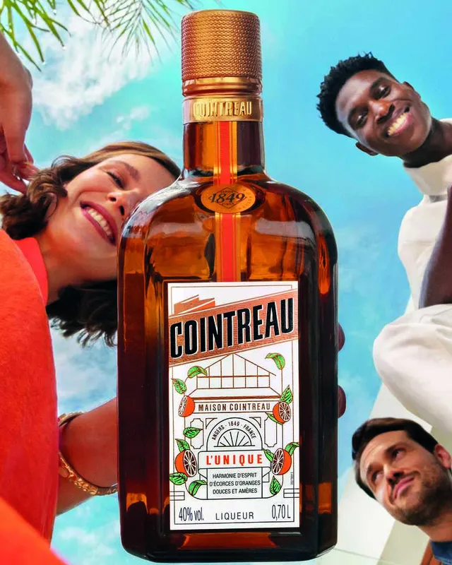 Cointreau bottle in the center with three people leaning in from above against a bright blue sky background.
