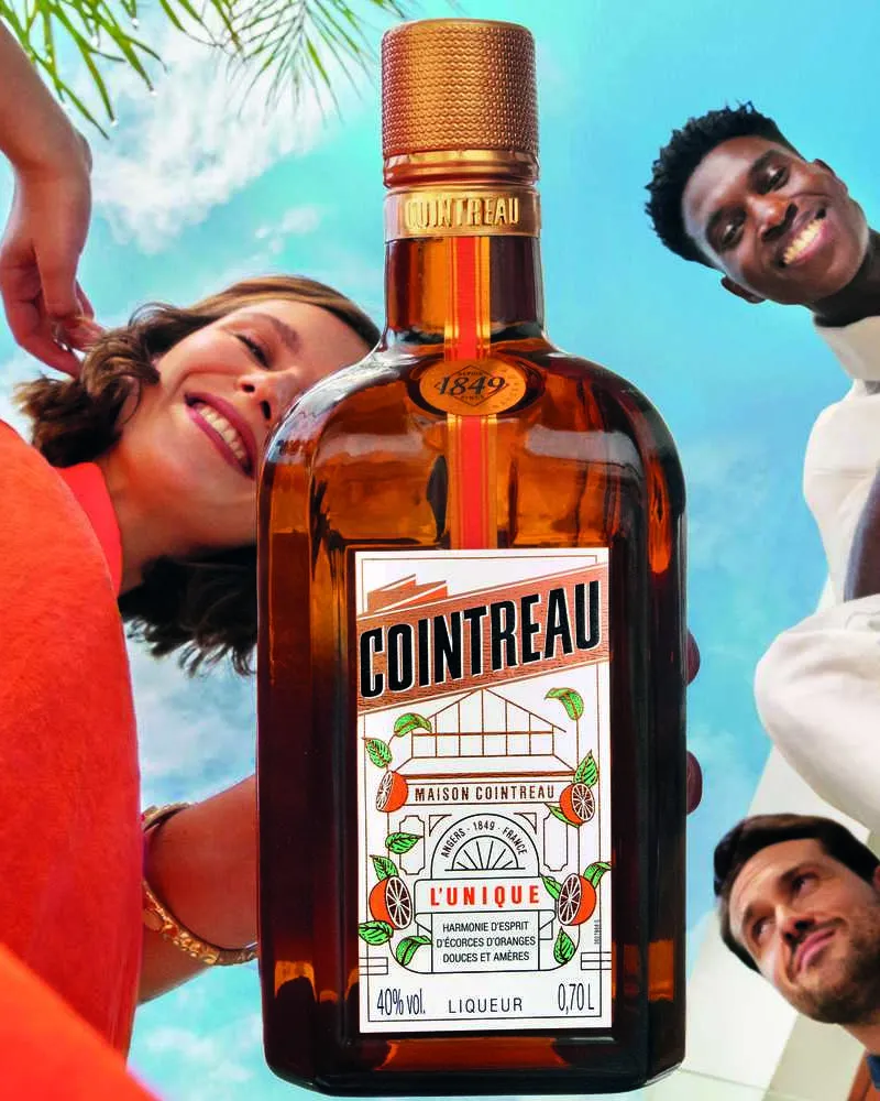 Cointreau bottle in the center with three people leaning in from above against a bright blue sky background.