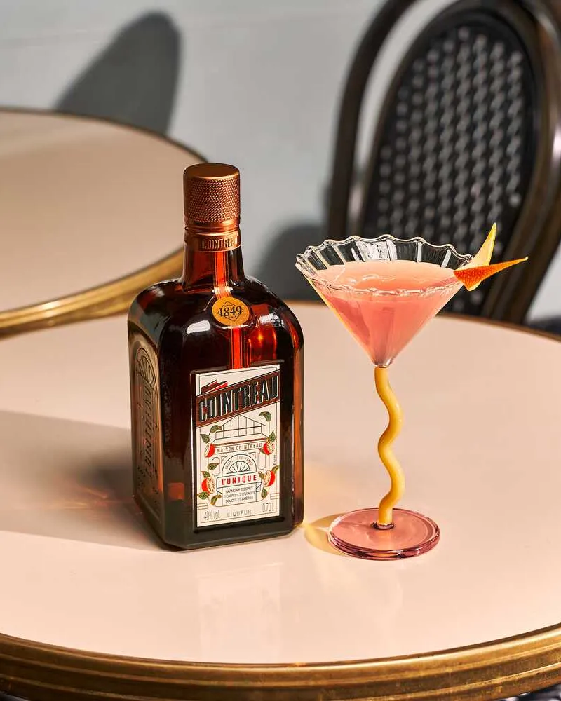 Cosmopolitan cocktail in a decorative stemmed glass with an orange twist garnish, served beside a bottle of Cointreau on a café table.