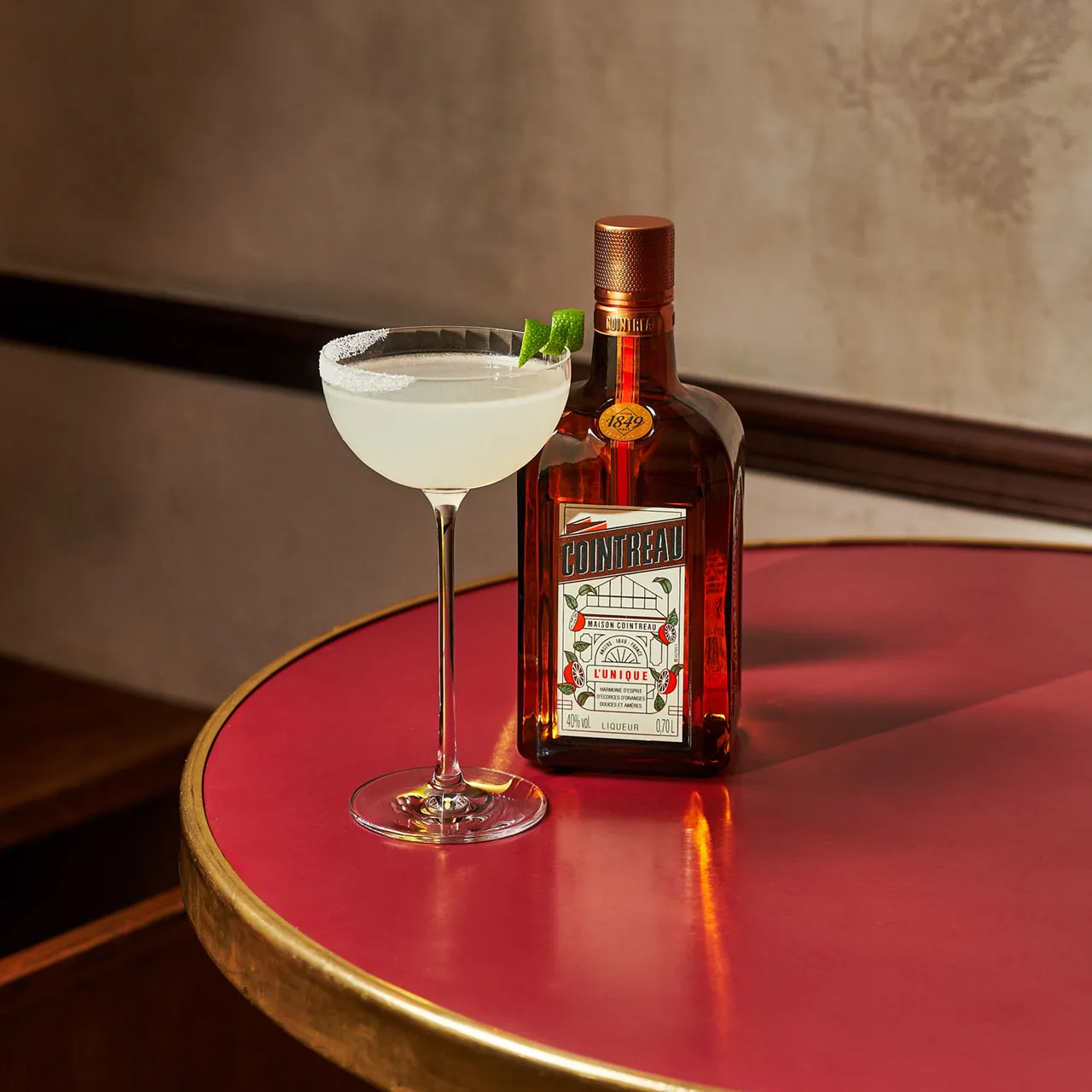 A bottle of Cointreau Orange liqueur beside a coupe with a pale yellow cocktail on a red table