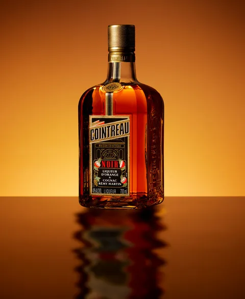 Side view of a Cointreau Noir bottle reflected on glossy surface with warm amber gradient backdrop