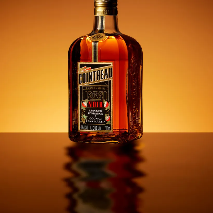 Side view of a Cointreau Noir bottle reflected on glossy surface with warm amber gradient backdrop