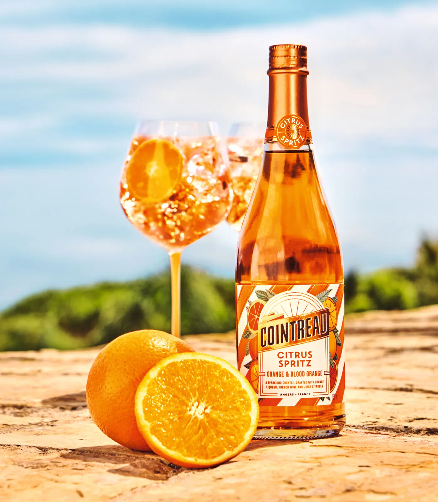 An arrangement of a bottle of Cointreau Citrus Spritz, a spritz glass and oranges in a summery setting