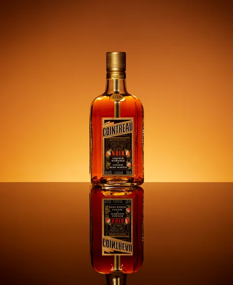 Cointreau Noir bottle reflected on glossy surface with warm amber gradient backdrop