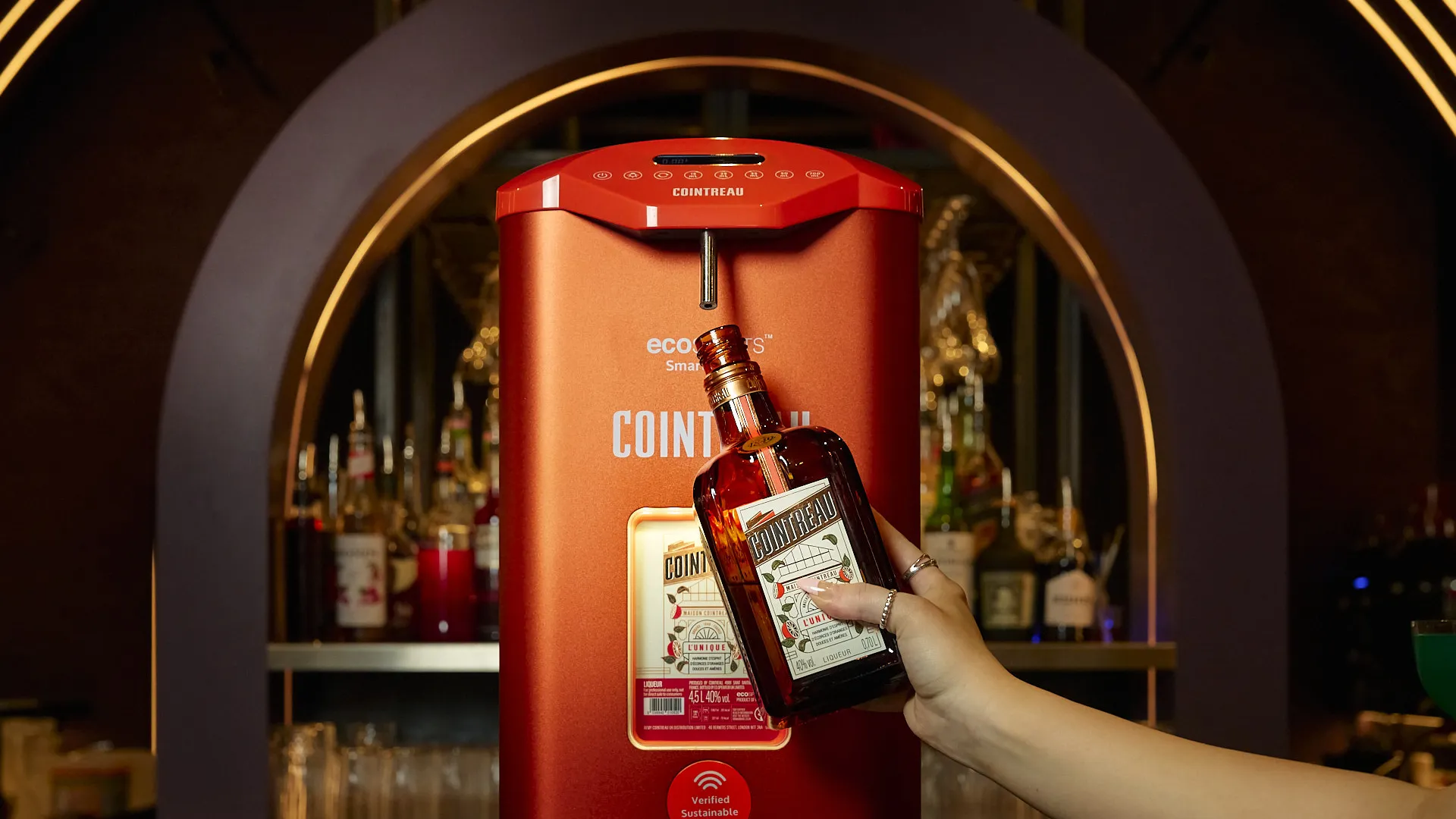 Cointreau SmartPour 2.0 refill station on a bar counter, with a hand holding a Cointreau bottle beneath the dispensing nozzle for refill.