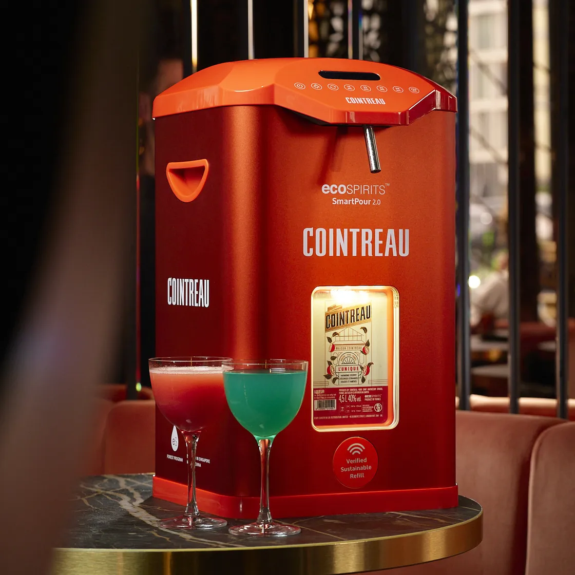 Cointreau SmartPour 2.0 refill station displayed on a marble table beside two colorful cocktails in a lounge-style bar setting.