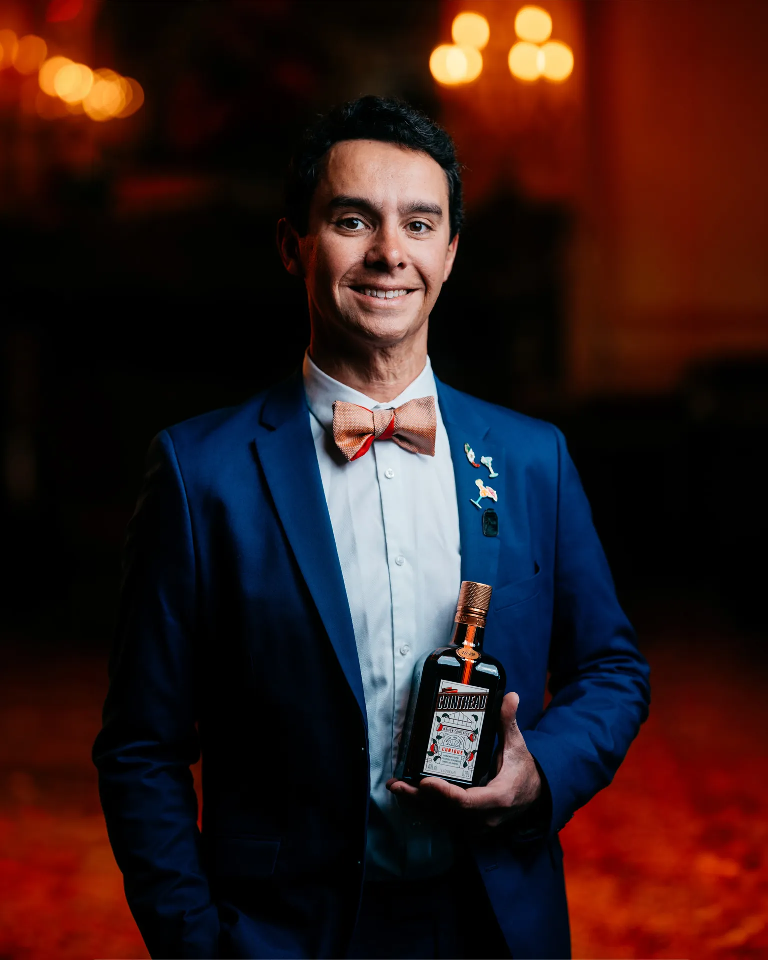 A medium shot of Alfred Cointreau holding a bottle of Cointreau Orange liqueur in front of a dark background with bokeh