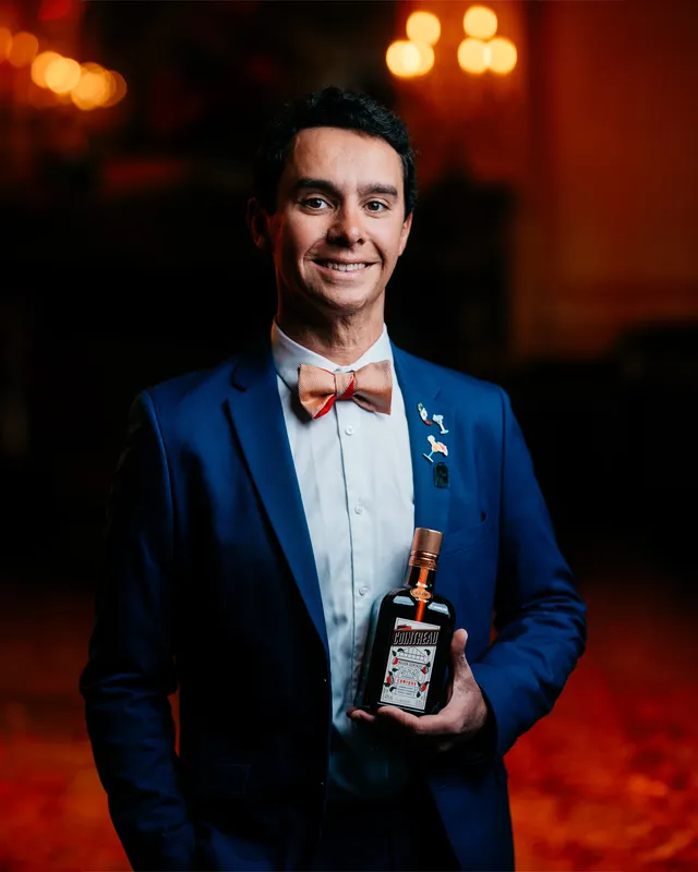 A medium shot of Alfred Cointreau holding a bottle of Cointreau Orange liqueur in front of a dark background with bokeh