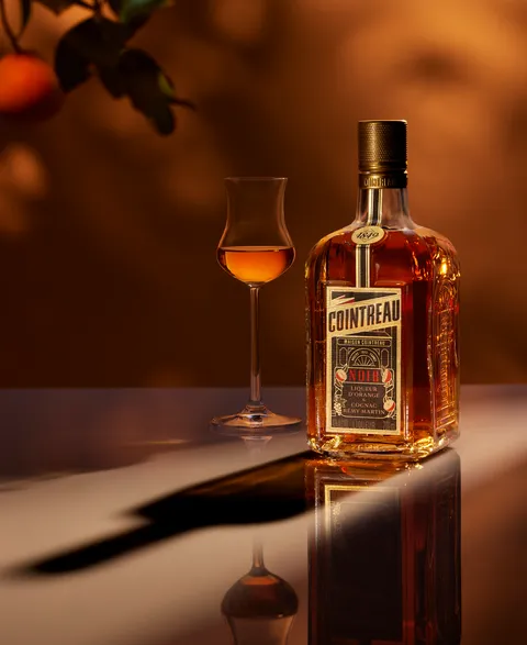 Cointreau Noir bottle reflected on glossy surface, amber backdrop with orange tree