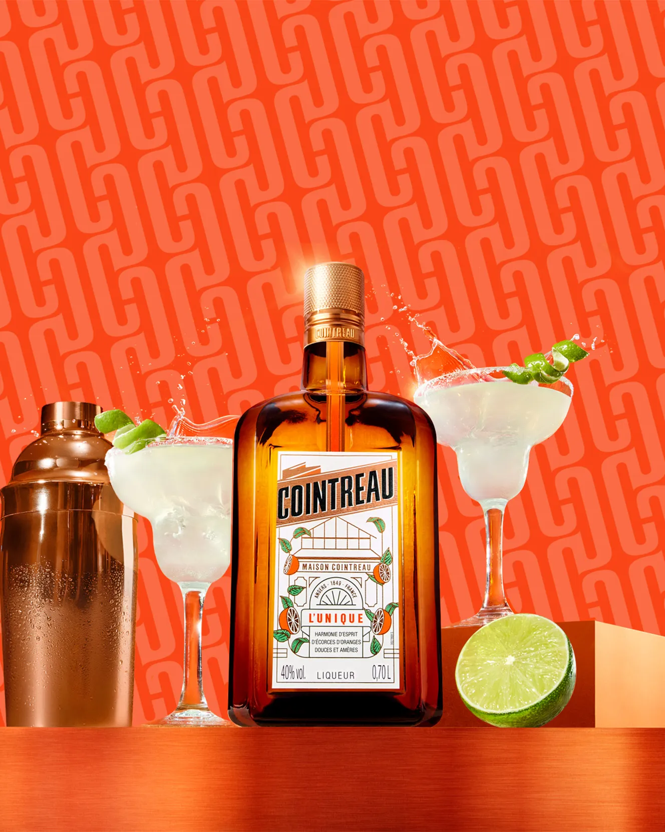 Cointreau bottle with margaritas, shaker, and lime on a vibrant orange background