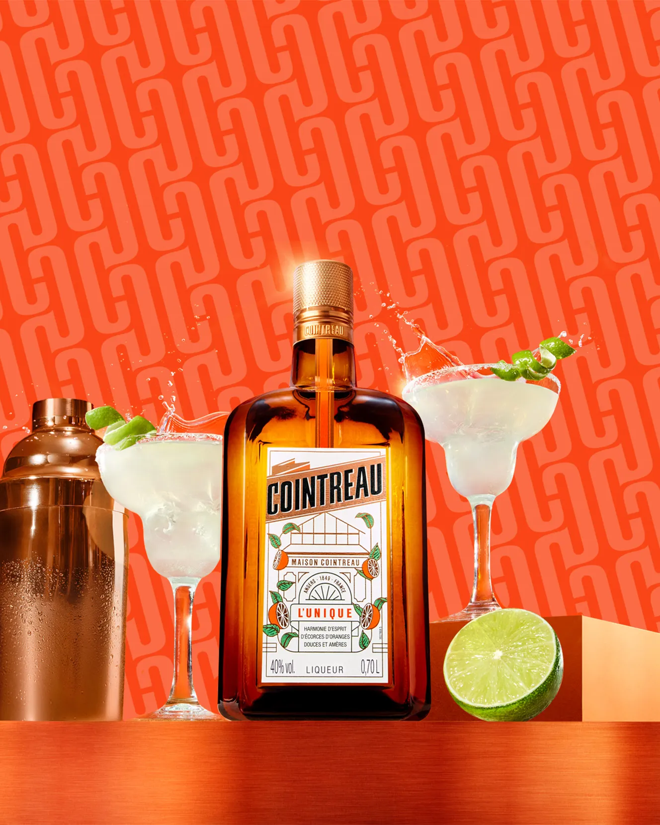 Cointreau bottle with margaritas, shaker, and lime on a vibrant orange background