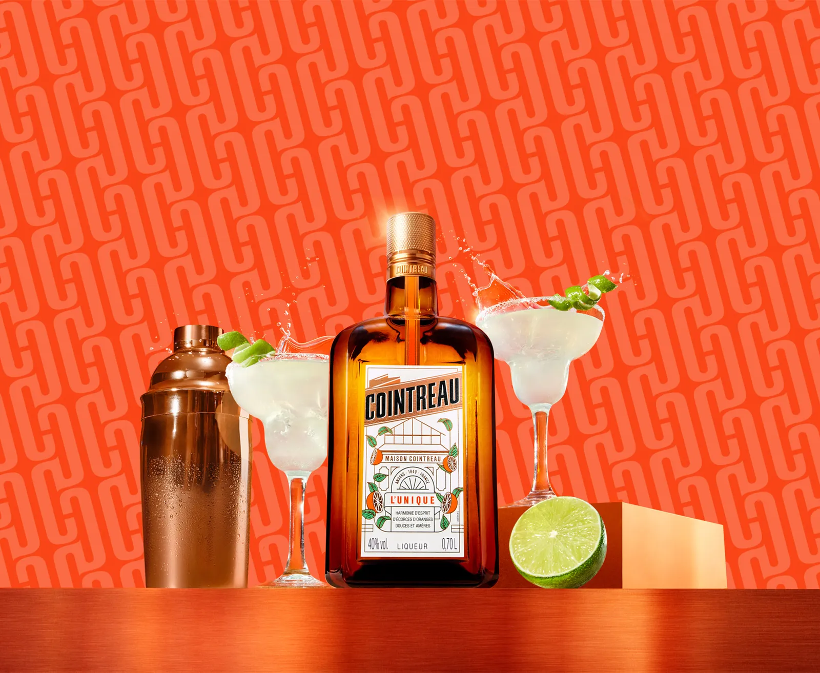 Cointreau bottle with margaritas, shaker, and lime on a vibrant orange background