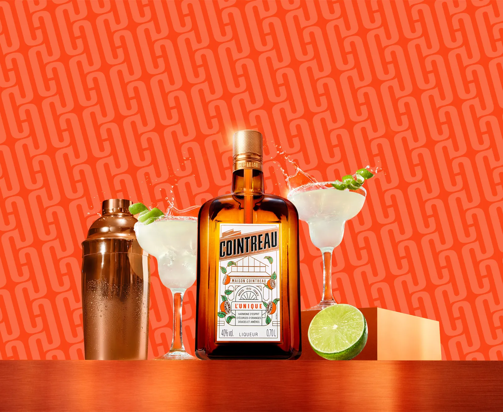 Cointreau bottle with margaritas, shaker, and lime on a vibrant orange background