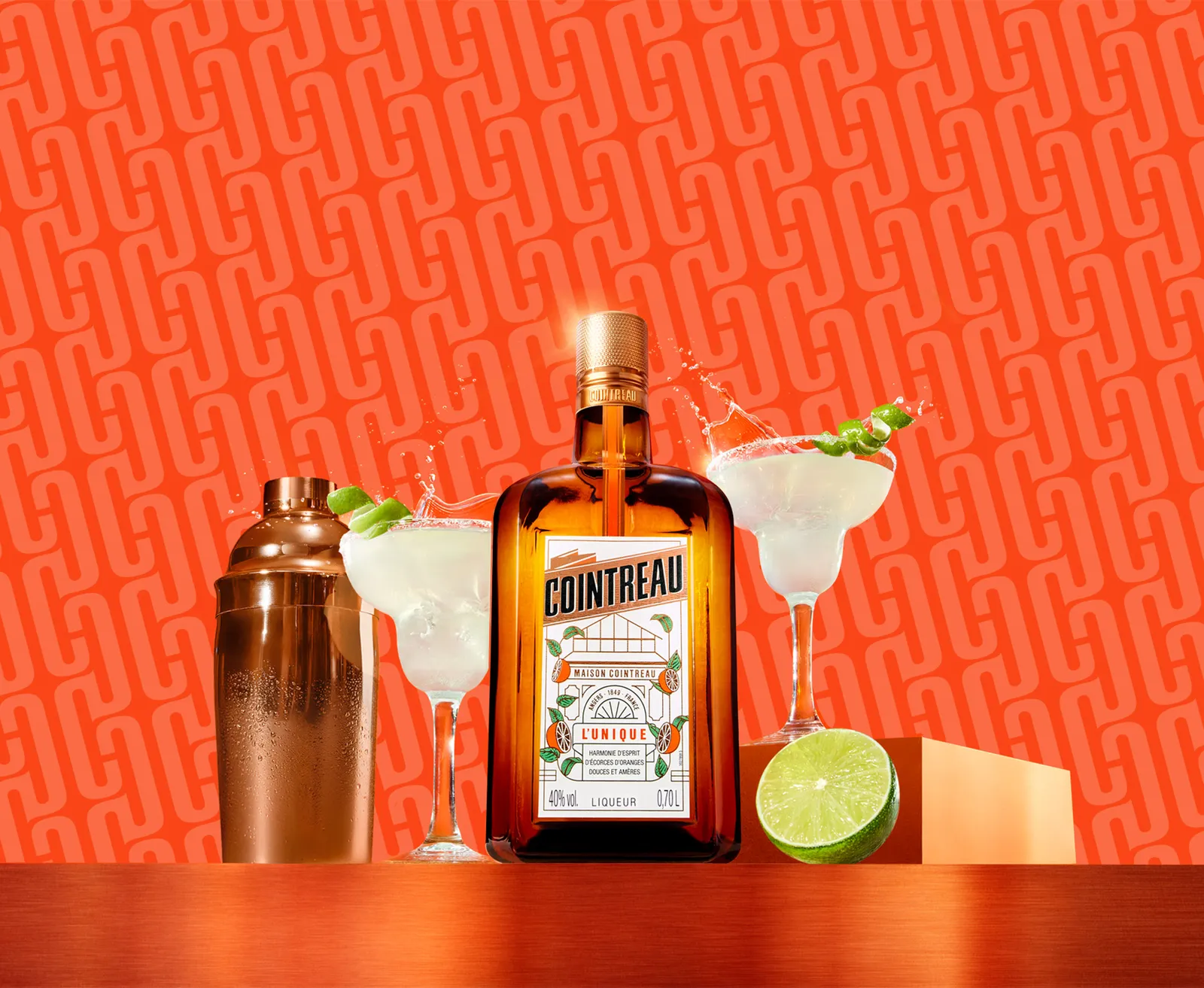 Cointreau bottle with margaritas, shaker, and lime on a vibrant orange background