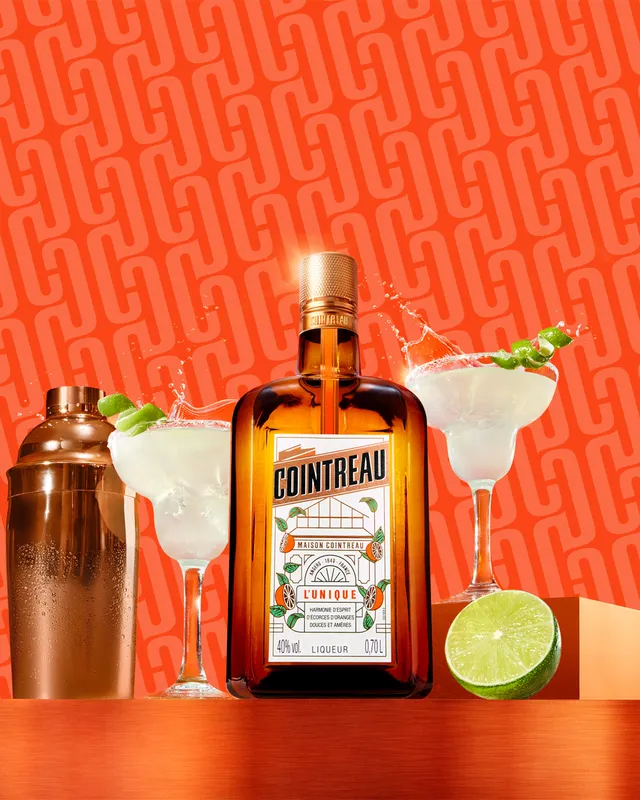 Cointreau bottle with margaritas, shaker, and lime on a vibrant orange background