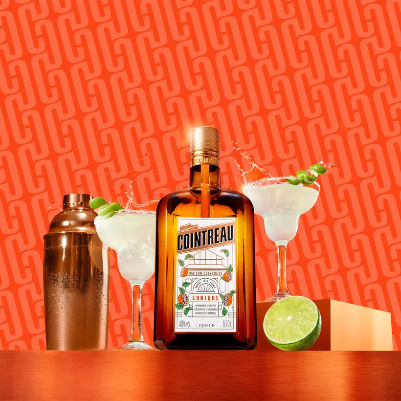 Cointreau bottle with margaritas, shaker, and lime on a vibrant orange background