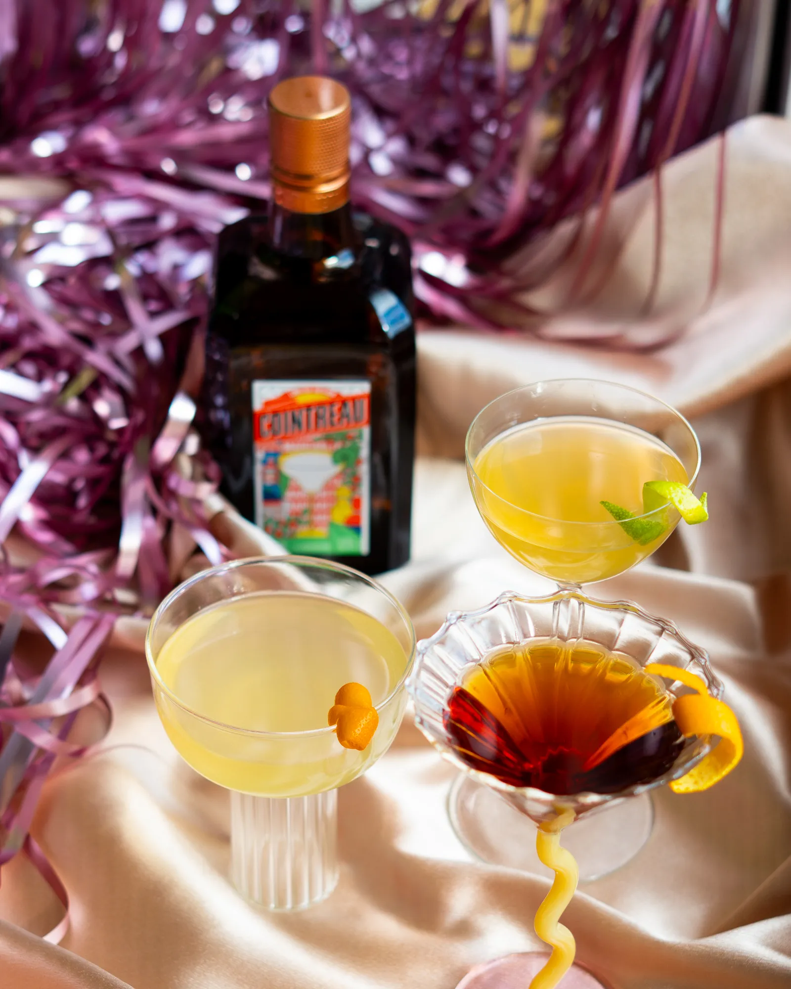 Three cocktails and a bottle of Cointreau arranged on satin fabric with pink metallic streamers in the background, creating a festive and elegant setting.
