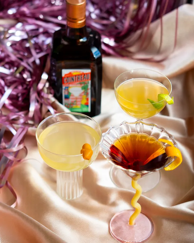 Three cocktails and a bottle of Cointreau arranged on satin fabric with pink metallic streamers in the background, creating a festive and elegant setting.
