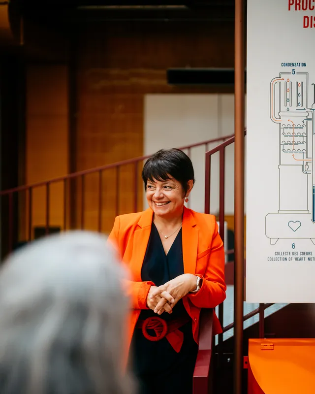 Person in a bright orange blazer standing beside an exhibition display panel and staircase in an indoor event space.