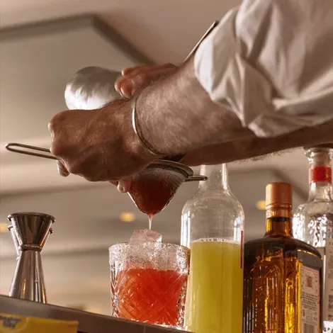 Mixologist straining red cocktail into ice-filled glass with bar tools and ingredients in background