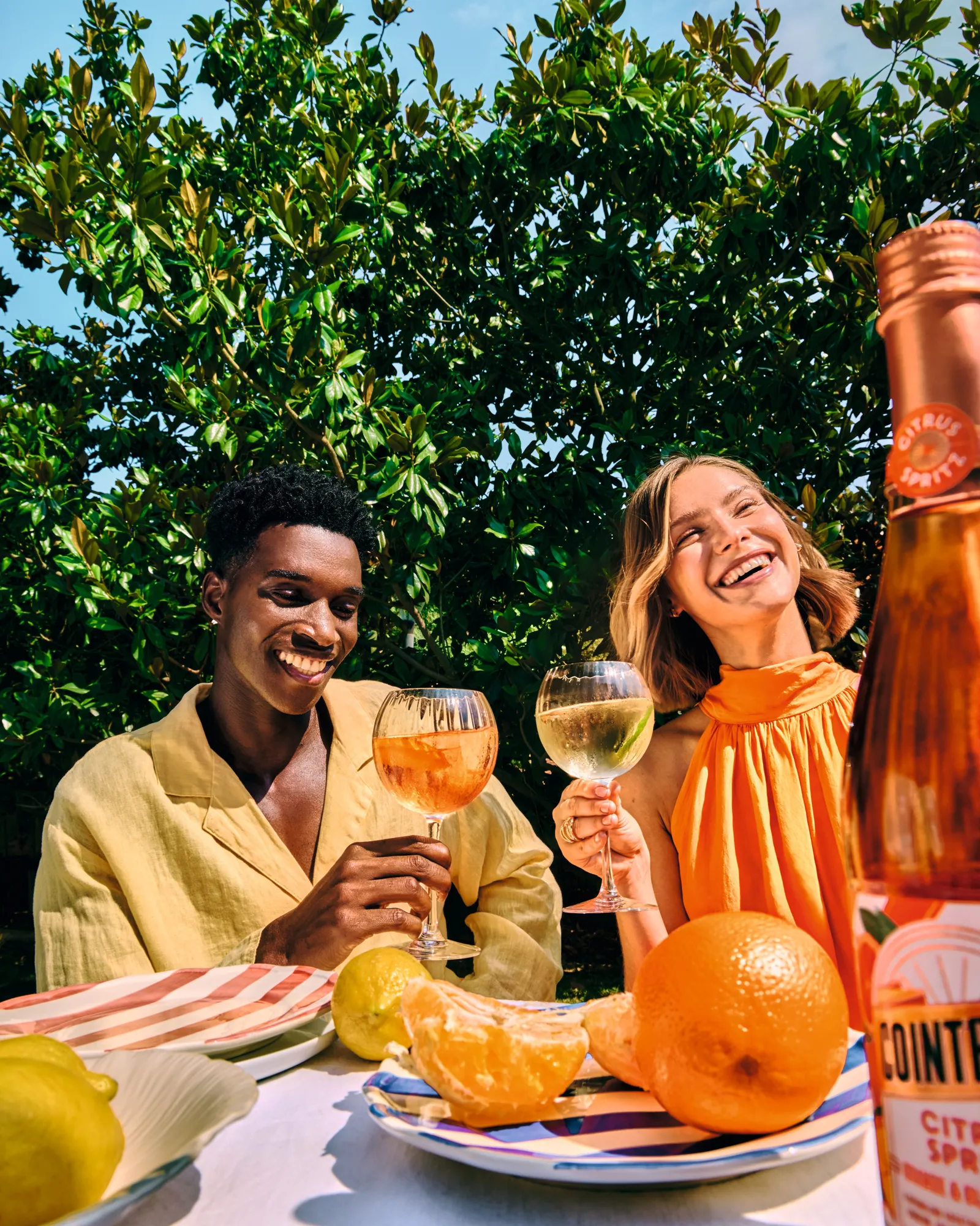 Two people enjoying citrus spritz drinks at outdoor table with fresh fruit and bottle