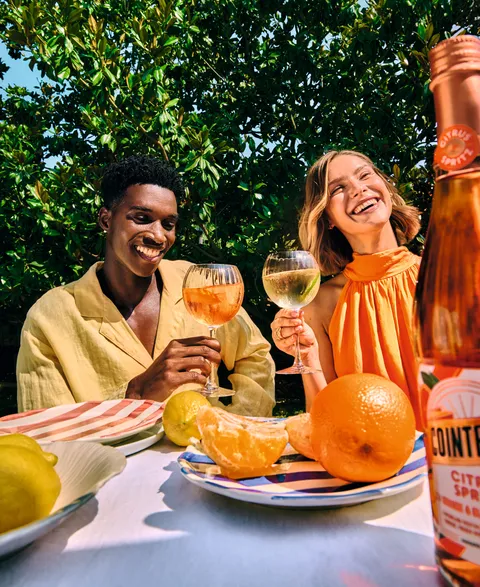 Two people enjoying citrus spritz drinks at outdoor table with fresh fruit and bottle