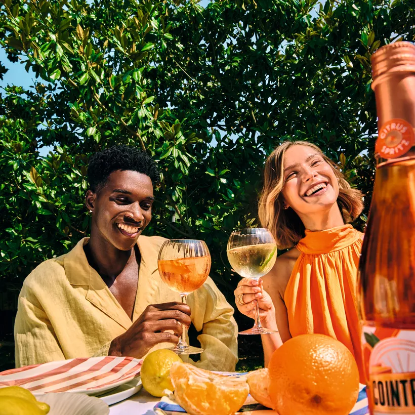 Two people enjoying citrus spritz drinks at outdoor table with fresh fruit and bottle