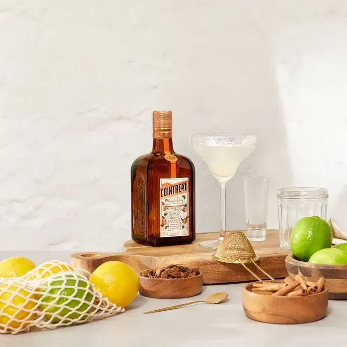Cointreau margarita setup with bottle, cocktail glass, limes and bar tools in front of a structured, white wall