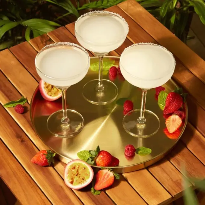 Three frozen margarita cocktails on a gold tray, garnished with fresh fruit and mint, surrounded by strawberries, raspberries, and passion fruit on a wooden table.