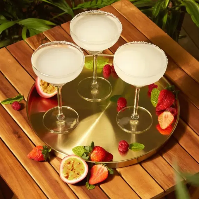 Three frozen margarita cocktails on a gold tray, garnished with fresh fruit and mint, surrounded by strawberries, raspberries, and passion fruit on a wooden table.
