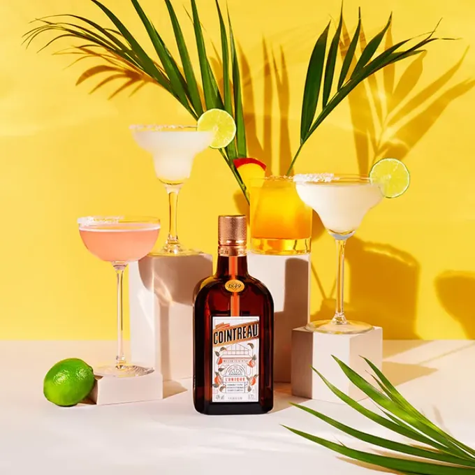 An arrangement of a bottle of Cointreau and several different Margarita cocktails in front of a yellow wall
