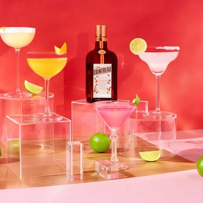 An arrangement of a bottle of Cointreau L’Unique, several cocktails and some limes in front of a hot pink wall
