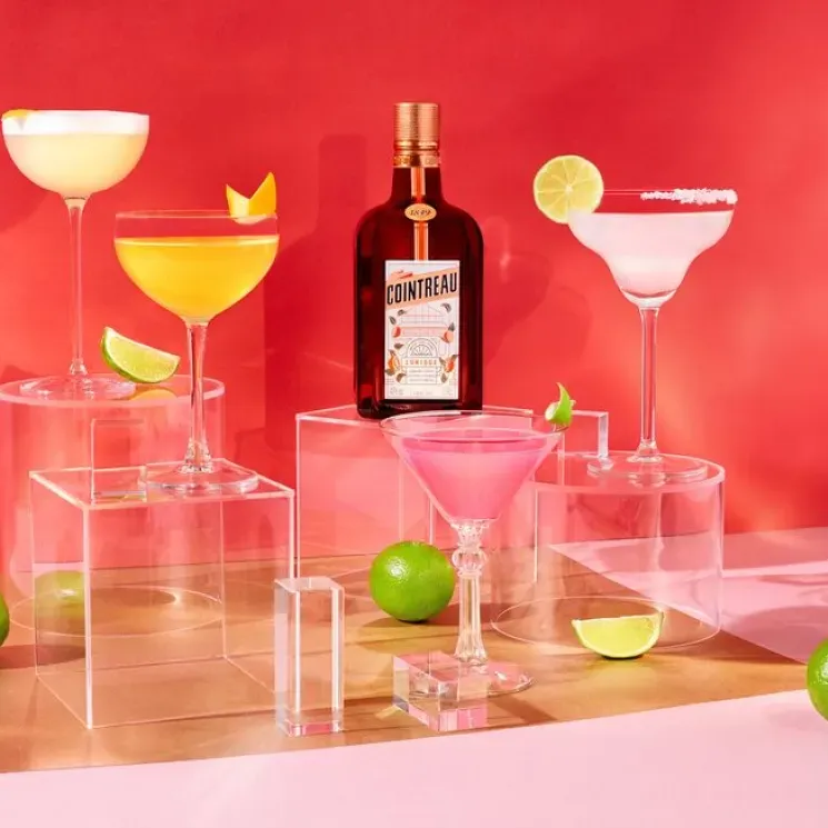 An arrangement of a bottle of Cointreau L’Unique, several cocktails and some limes in front of a hot pink wall