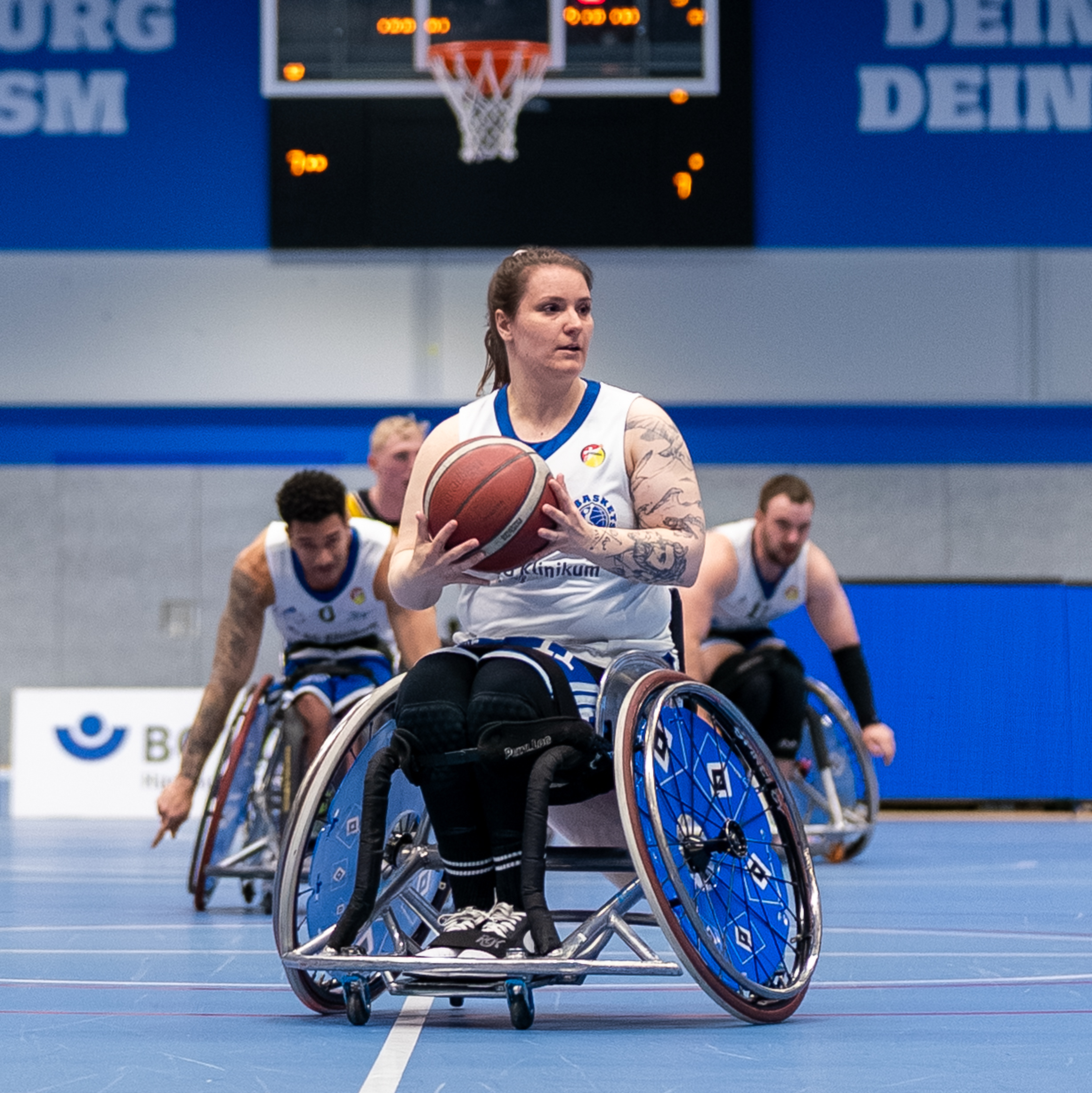 A female wheelchair basketball player with tattoos holds the ball, teammates behind, on an indoor court near a basketball hoop.