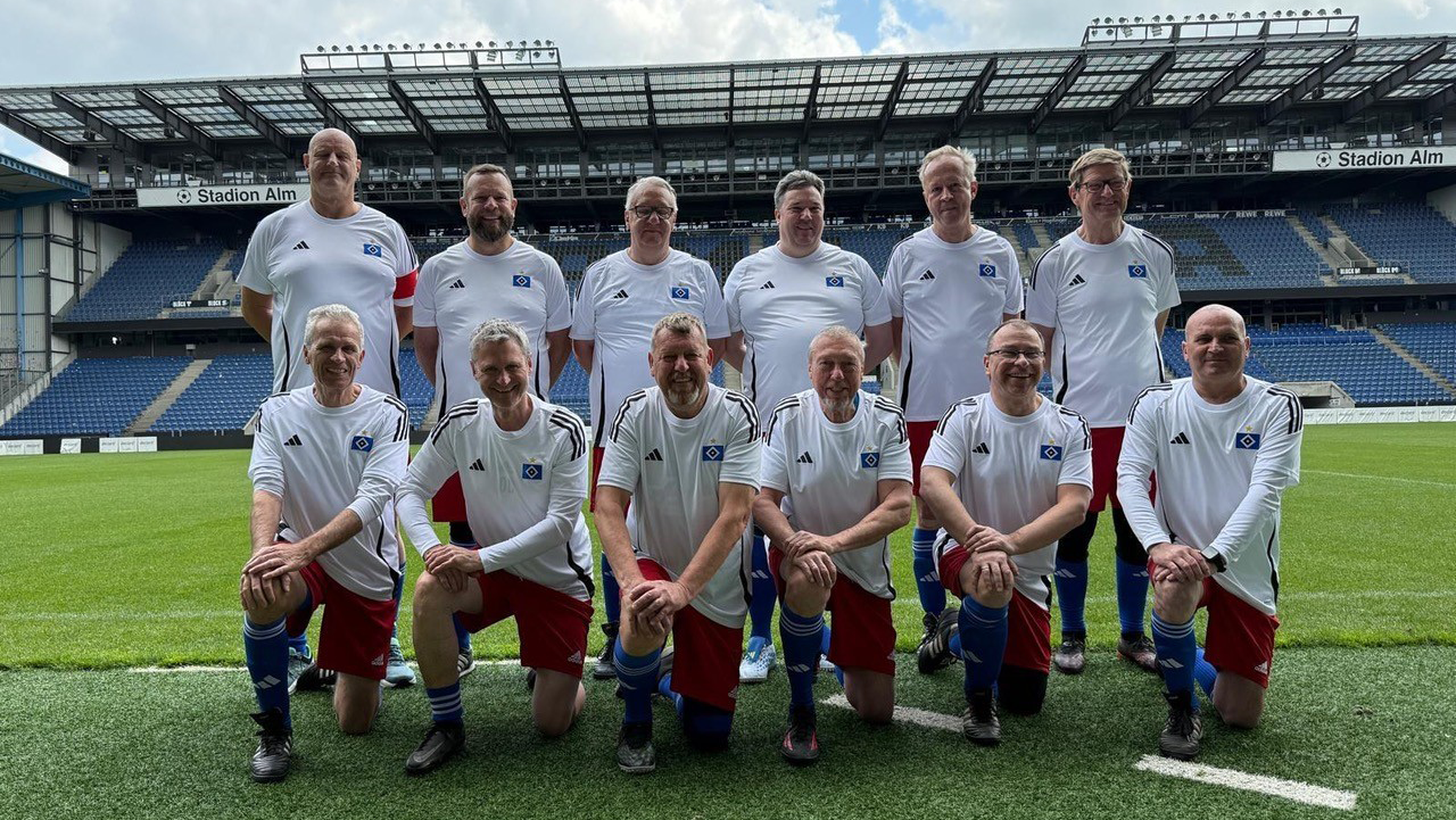 A football team of twelve men in white jerseys and red shorts posing on a stadium field with empty blue stands in the background.