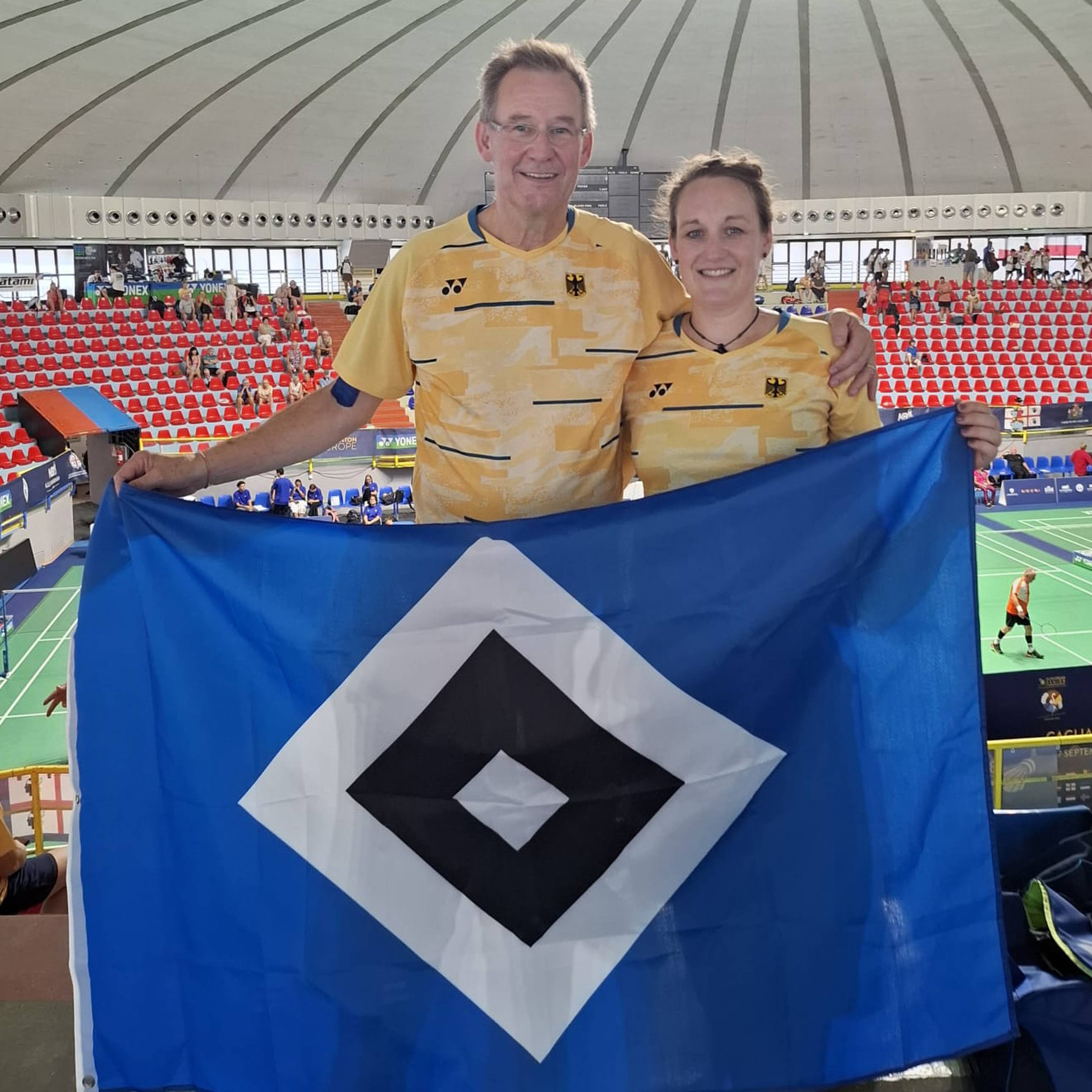 Two people in matching yellow shirts hold a blue flag with a black and white diamond pattern at an indoor sports venue.