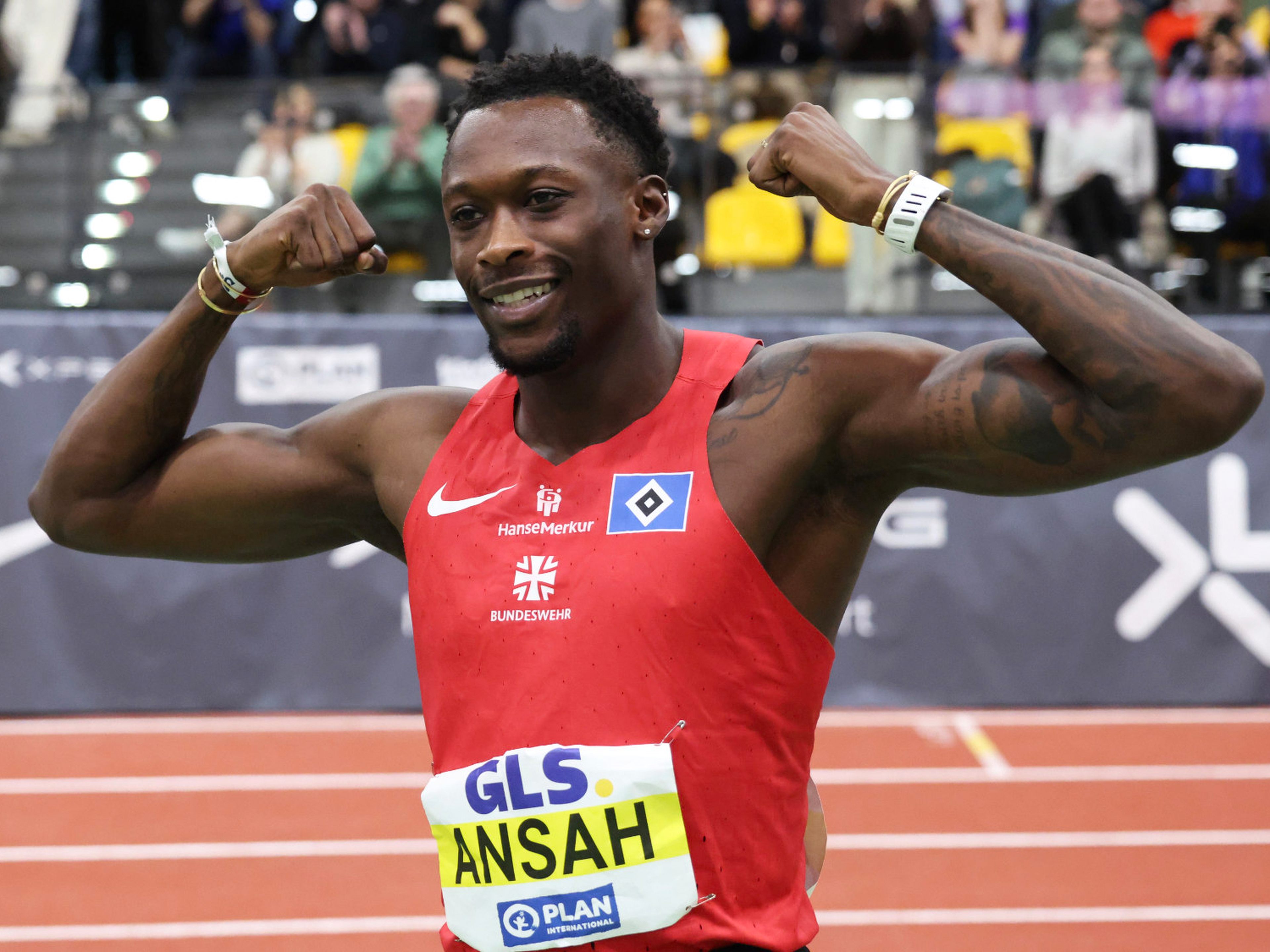 Athlete in a red jersey celebrates by flexing muscles on a track, wearing a race bib labeled "ANSAH," with a cheering crowd in the background.